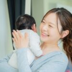 赤ちゃんを抱っこしながら優しい笑顔で立つママの様子。疲れを感じさせない健康的な姿勢が印象的な写真。