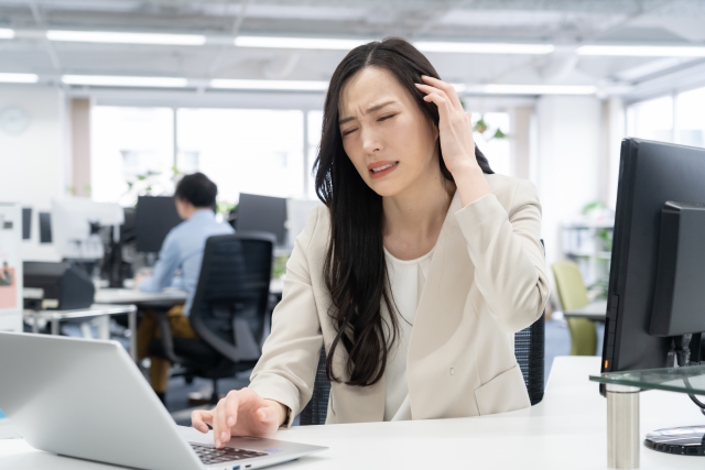 パソコン作業中に頭を押さえ、肩こりからくる頭痛に悩む女性