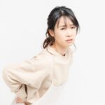 産後の腰痛に悩む女性が、エプロン姿で腰を押さえながら辛そうな表情を浮かべている様子。骨盤のゆがみや骨盤底筋群の弱化による負担をイメージした写真。