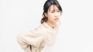 産後の腰痛に悩む女性が、エプロン姿で腰を押さえながら辛そうな表情を浮かべている様子。骨盤のゆがみや骨盤底筋群の弱化による負担をイメージした写真。
